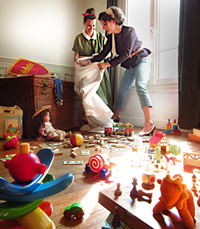 Photo. Le parquet de la pièce est rempli de jouet d'enfants, en arrière plan deux femmes hilare tiennent un immense sac blanc.